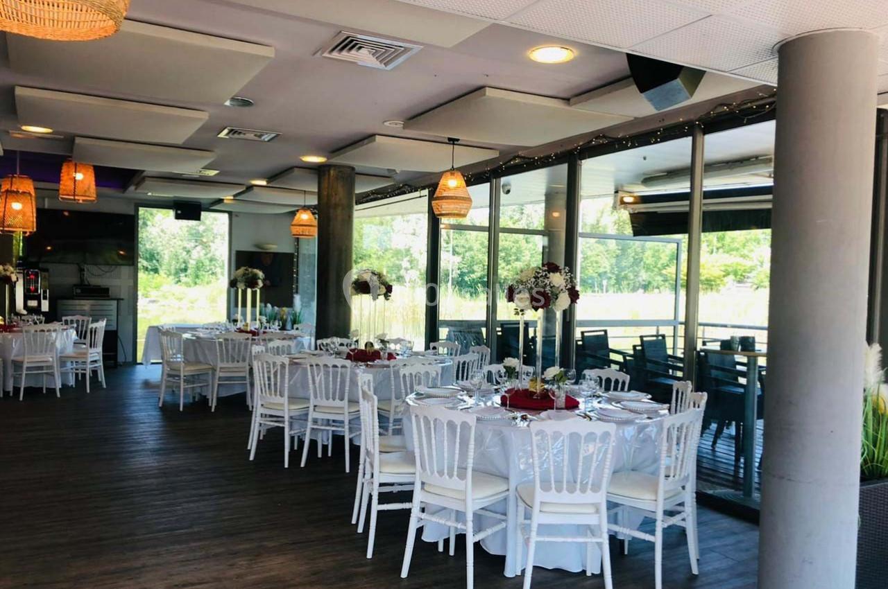 Salle de réception lumineuse avec tables dressées, chaises blanches et grandes baies vitrées donnant sur un espace vert.