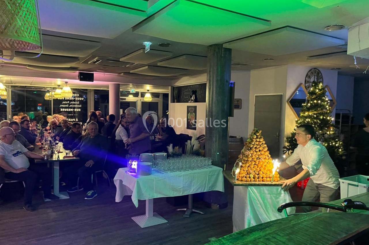 Un serveur présente une pièce montée éclairée dans un restaurant animé, décoré pour les fêtes avec un sapin de Noël.
