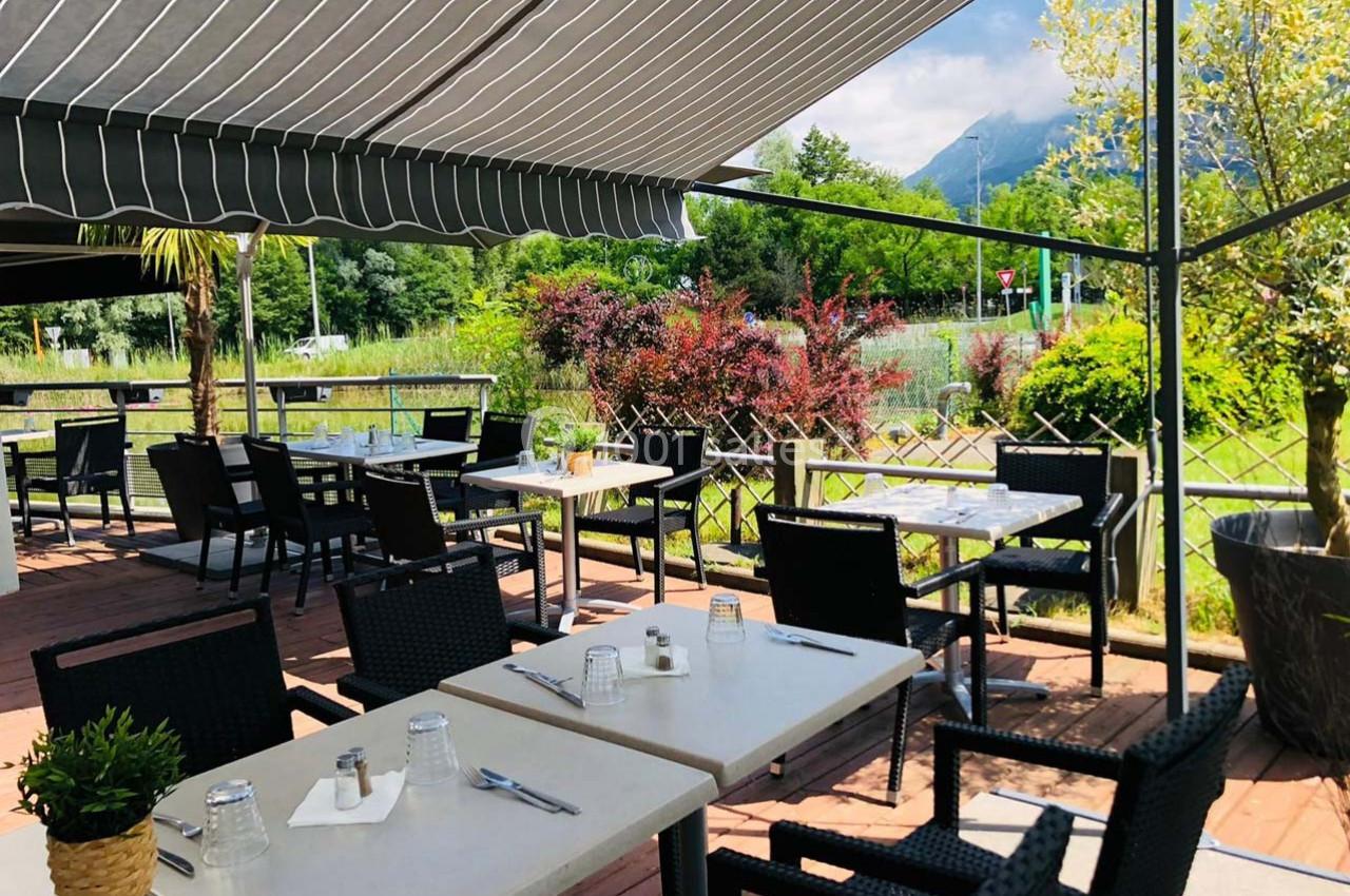 Terrasse extérieure avec tables et chaises sous un store, entourée de verdure et offrant une vue sur des montagnes.