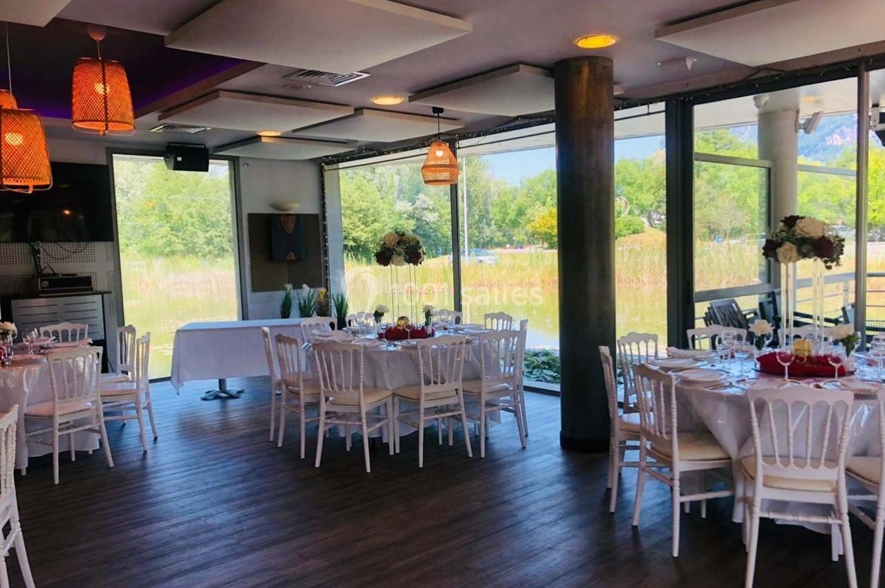 Salle lumineuse avec tables dressées pour un repas, grandes baies vitrées donnant sur un paysage verdoyant.