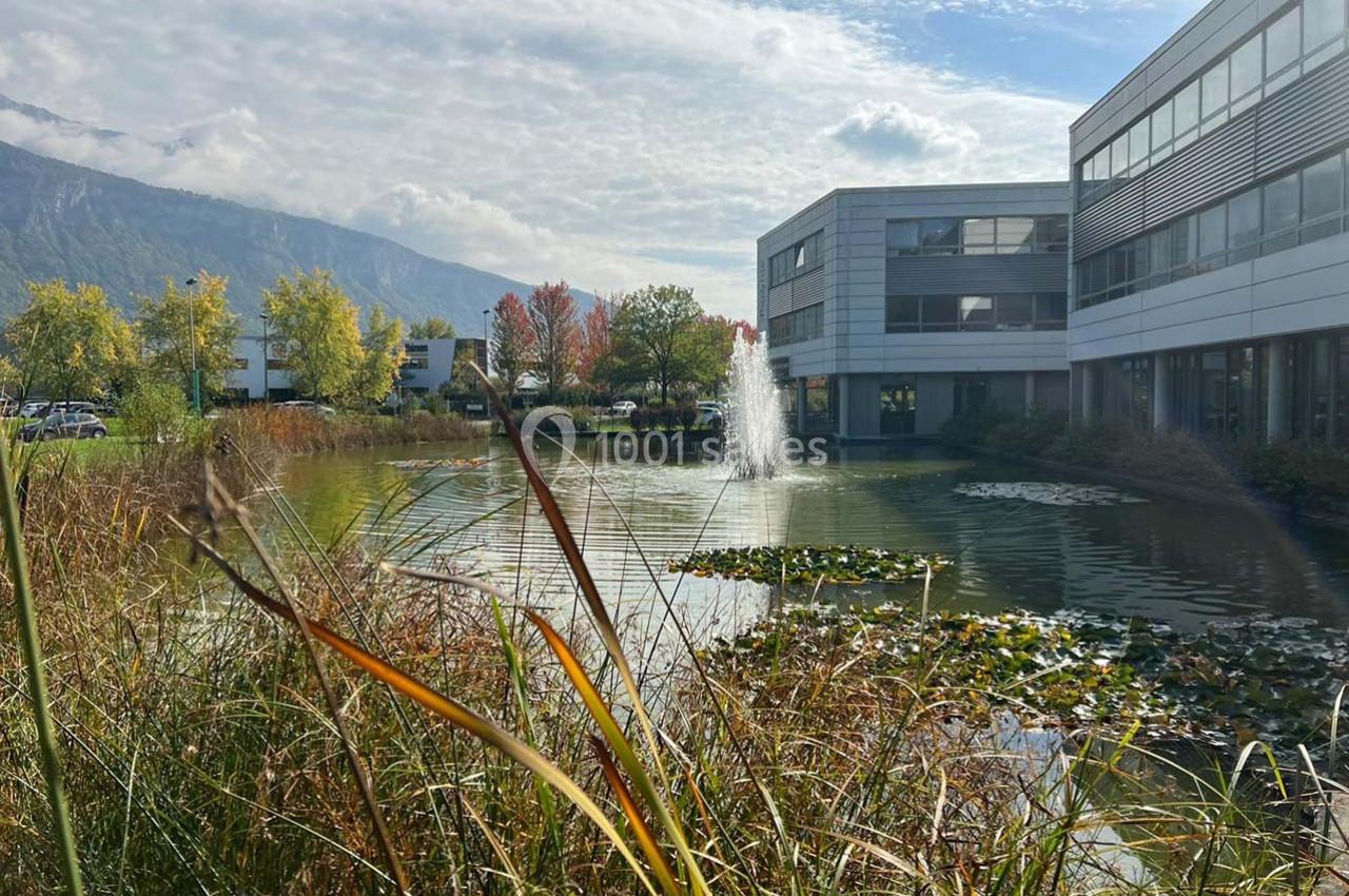 Étang entouré de végétation avec des nénuphars, une fontaine et des bâtiments modernes en arrière-plan.
