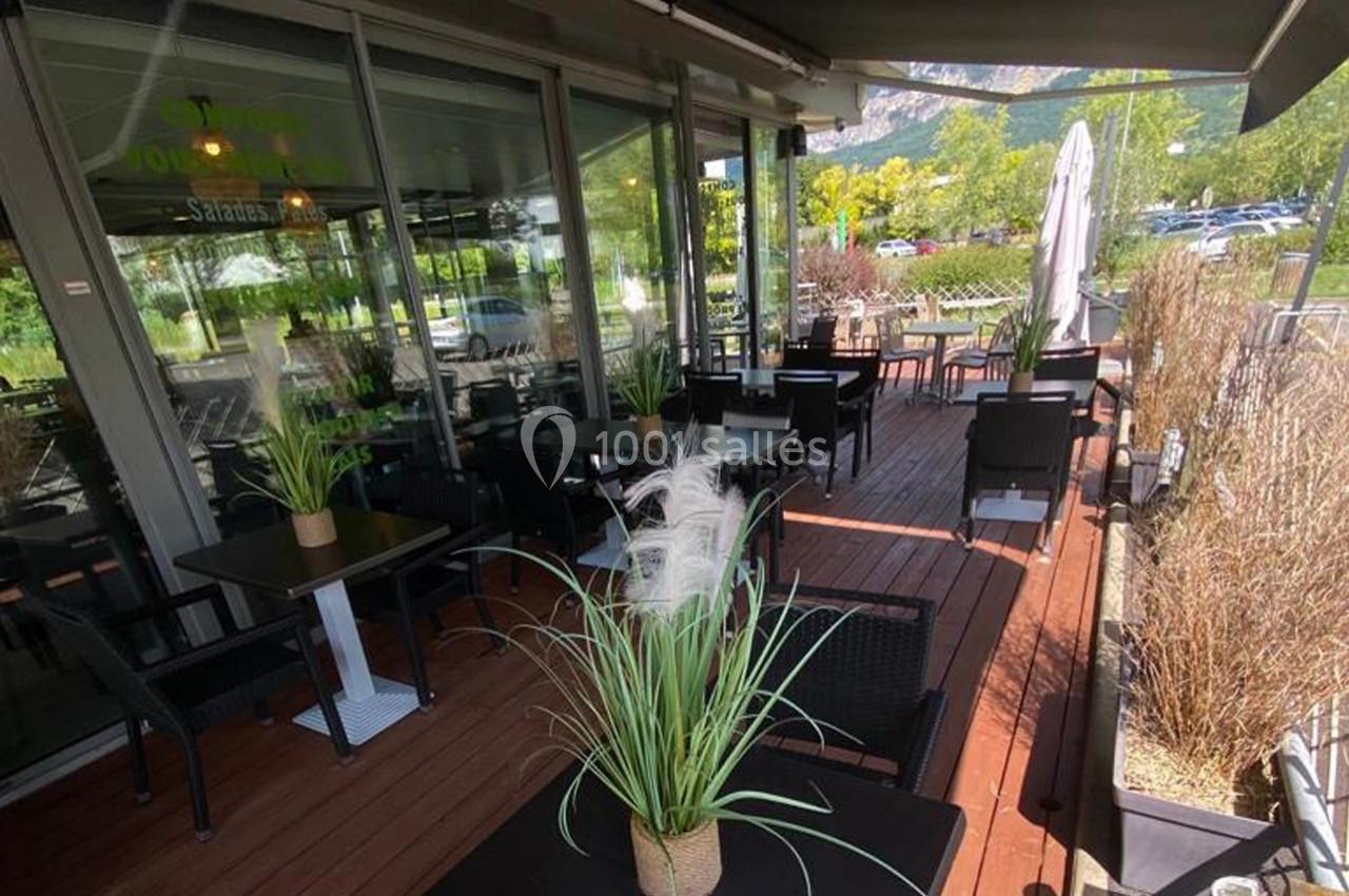 Terrasse en bois avec tables et chaises noires, entourée de plantes, donnant sur un espace extérieur verdoyant.