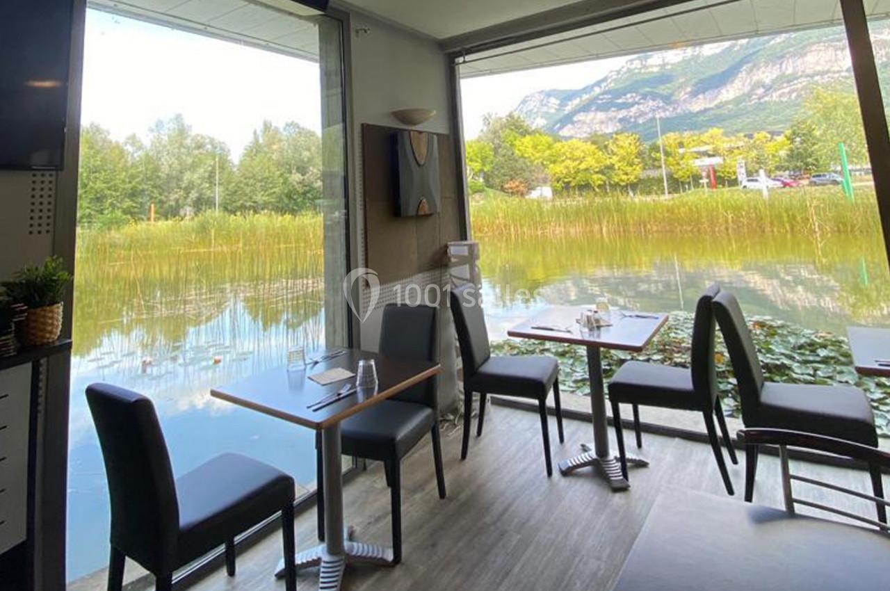 Salle de restaurant avec baies vitrées donnant sur un étang bordé de végétation et des montagnes en arrière-plan.