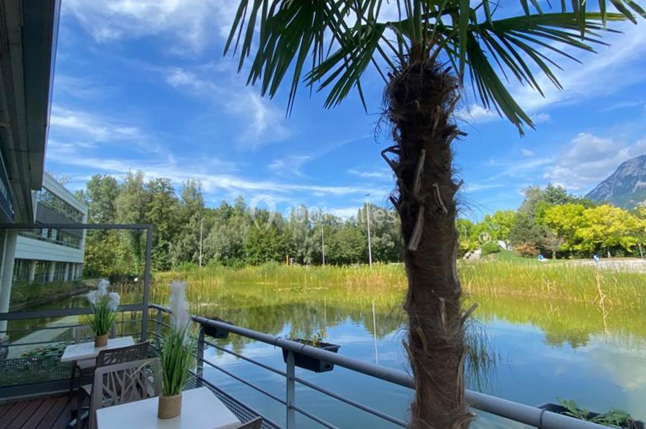 Vue d'une terrasse avec tables et plantes, donnant sur un étang entouré de végétation et montagnes sous un ciel bleu.