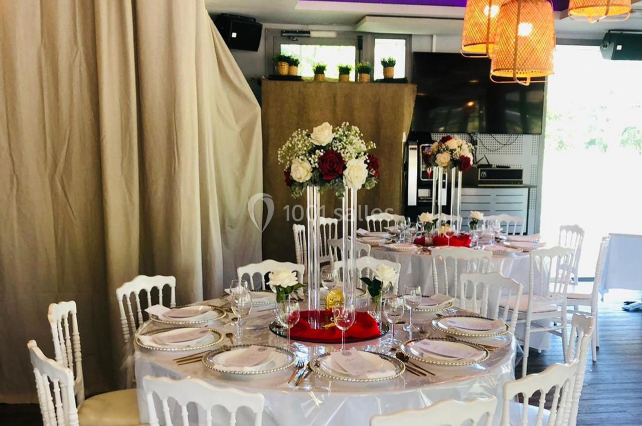 Salle décorée pour un événement avec tables rondes dressées, chaises blanches et centres de table floraux.