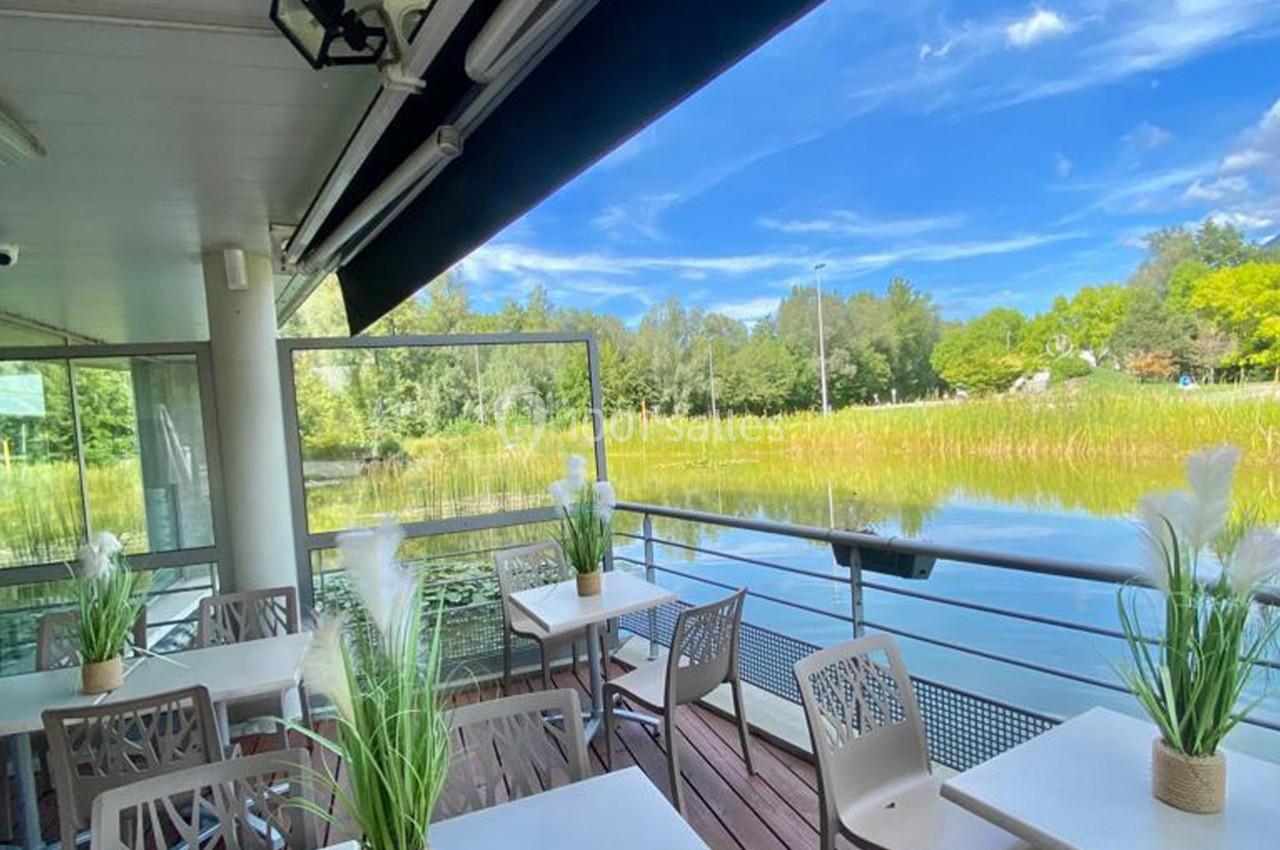 Terrasse avec tables et chaises donnant sur un plan d'eau entouré de végétation sous un ciel bleu.