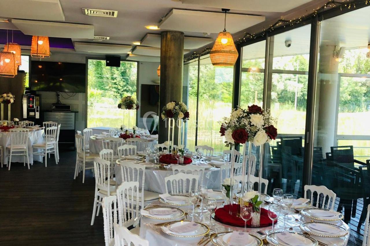 Salle de réception lumineuse avec tables rondes décorées de fleurs rouges et blanches, vue sur un extérieur verdoyant.