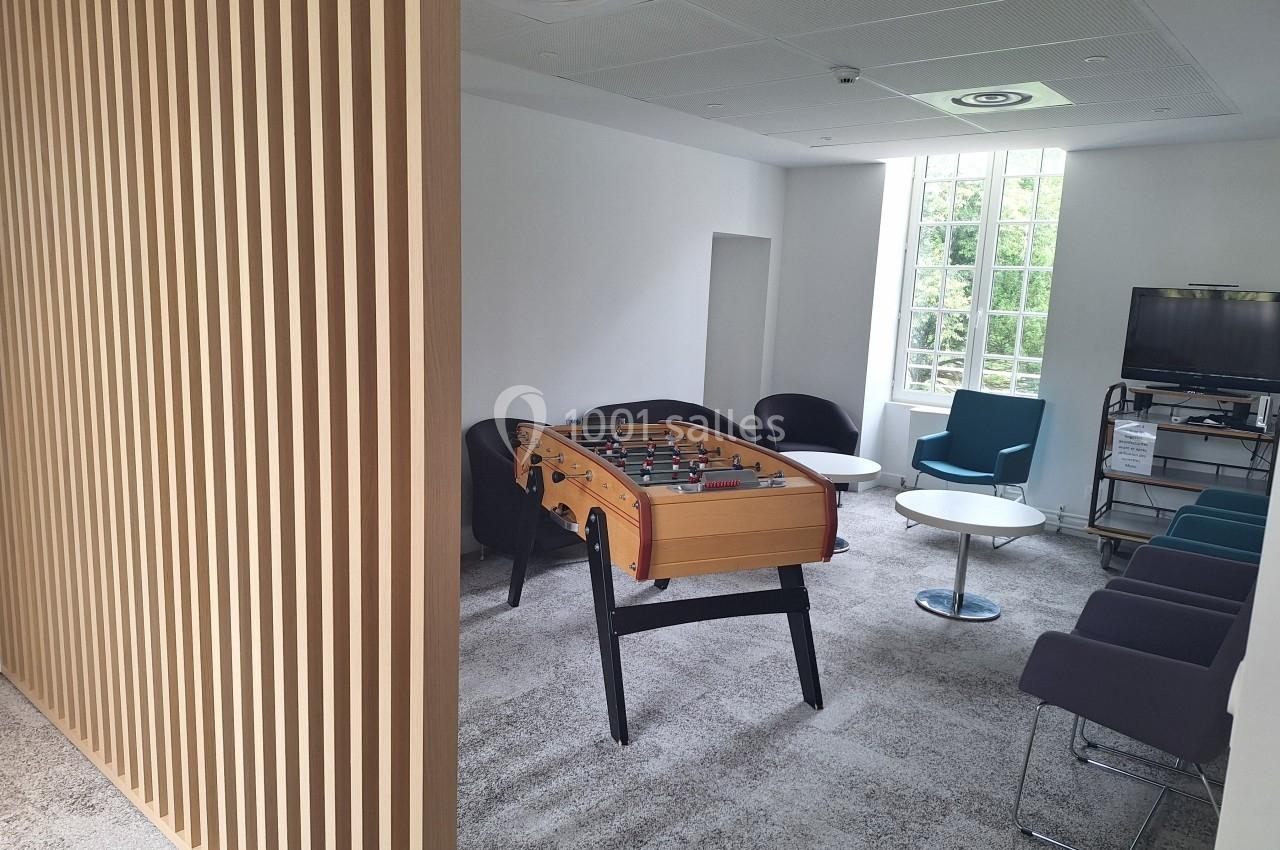 Salle de détente avec baby-foot, fauteuils, tables basses et télévision, éclairée par une grande fenêtre.