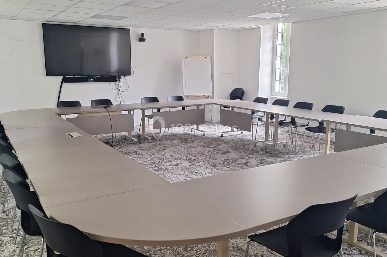 Salle de réunion lumineuse avec tables disposées en U, chaises noires, écran mural et tableau blanc.