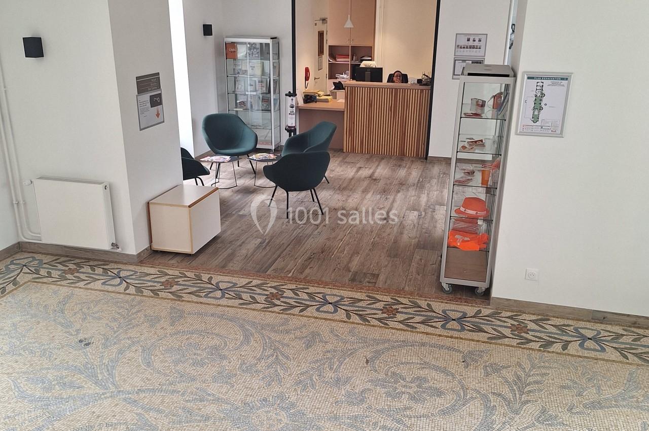 Espace d'accueil lumineux avec sol en mosaïque, fauteuils, comptoir en bois et vitrines d'exposition.