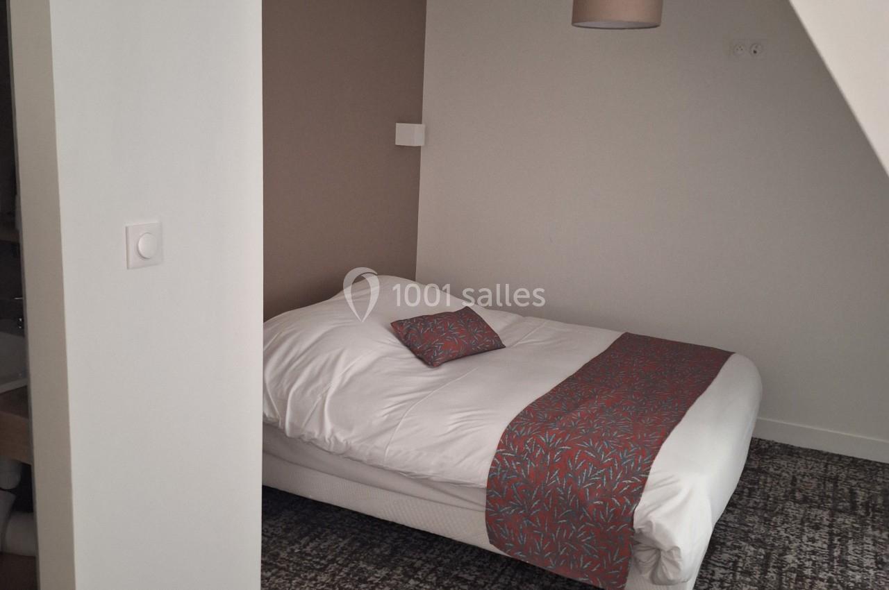 Lit double avec couverture et coussin décoratifs rouges dans une chambre aux tons neutres, éclairée par une suspension.