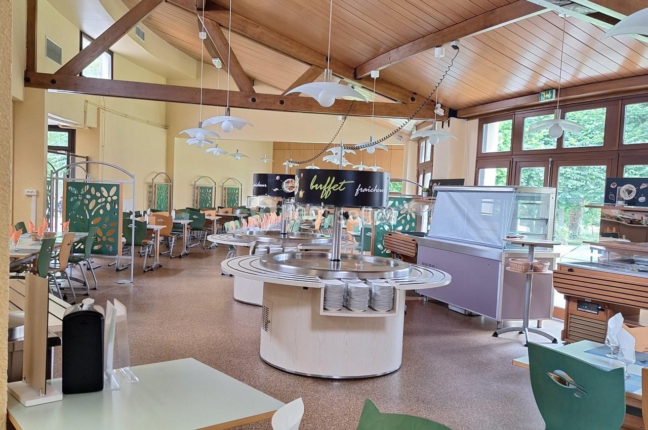 Salle de restaurant lumineuse avec tables dressées, buffet central et mobilier en bois sous un plafond en poutres apparentes.