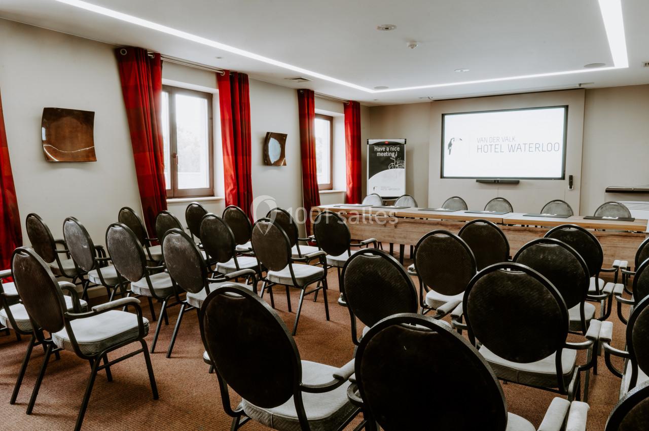 Salle de conférence avec rangées de chaises noires, écran de projection et rideaux rouges aux fenêtres.