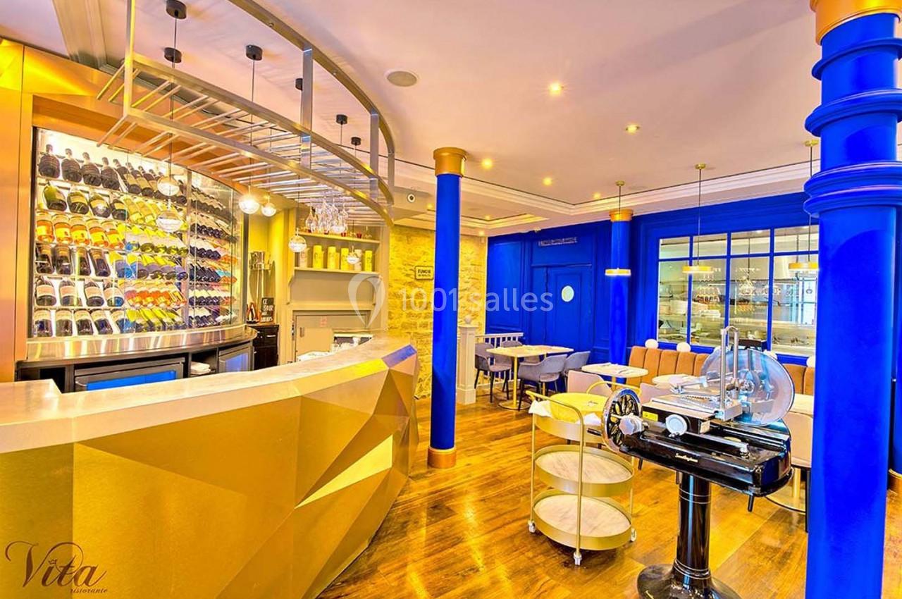 Intérieur d'un restaurant avec comptoir doré, étagères à bouteilles, tables modernes et murs aux tons bleus et jaunes.