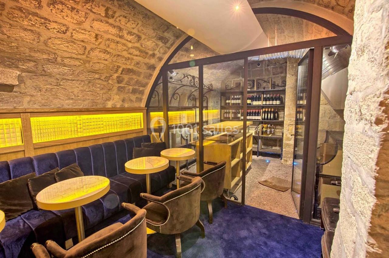 Salle voûtée en pierre avec tables, fauteuils en velours et étagères de bouteilles de vin en arrière-plan.