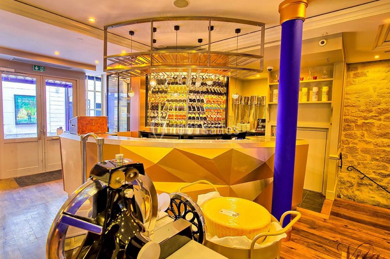 Intérieur d'une boutique avec un comptoir doré, une cave à vin vitrée, une trancheuse et une meule de fromage.