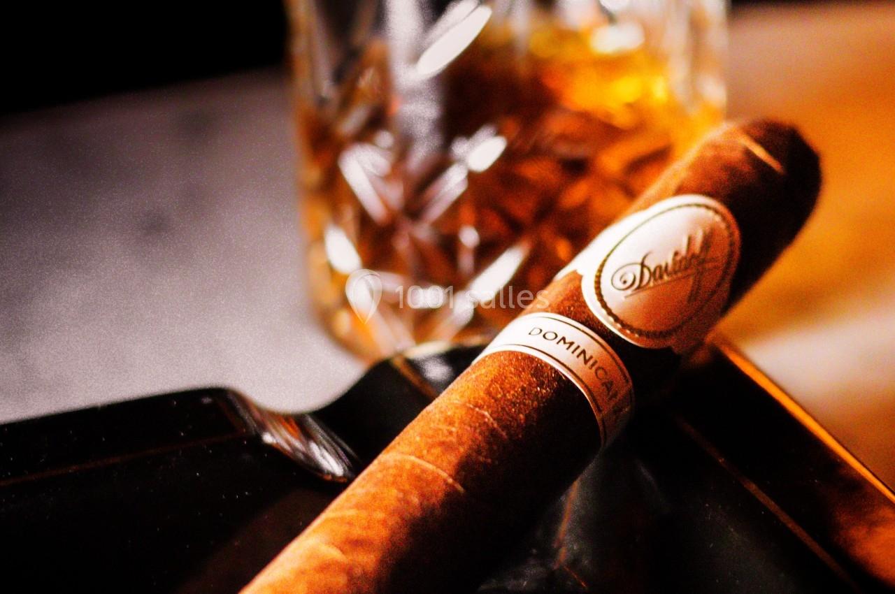 Un cigare posé sur un cendrier noir avec un verre de whisky ambré en arrière-plan.