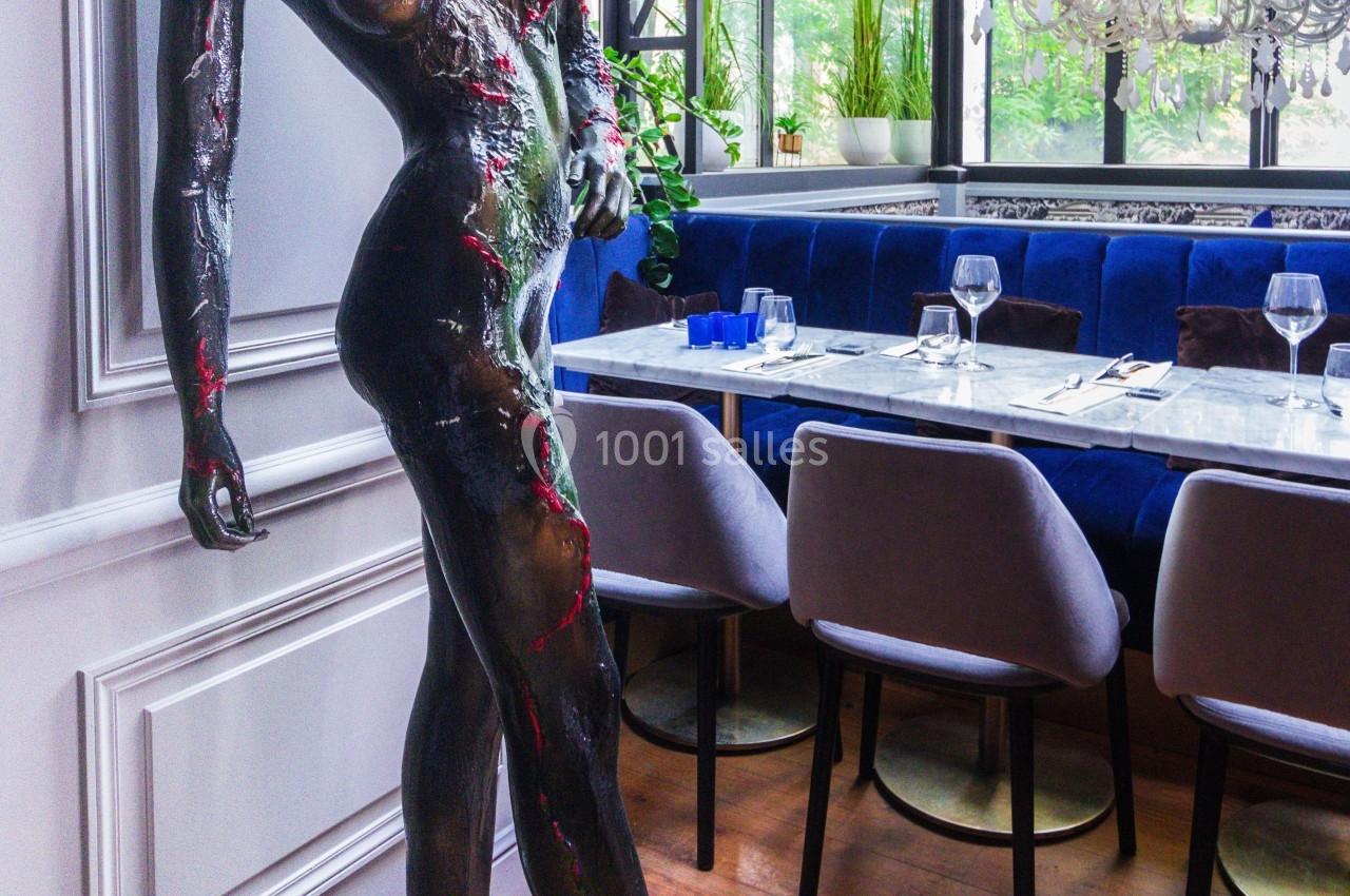 Statue moderne d'une silhouette humaine inclinée, exposée dans un restaurant lumineux avec des tables dressées.