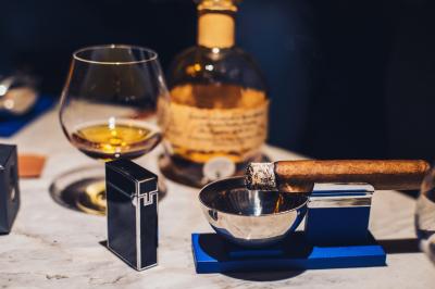 Verre de cognac, bouteille en arrière-plan, cigare allumé posé sur un cendrier et briquet sur une table en marbre.