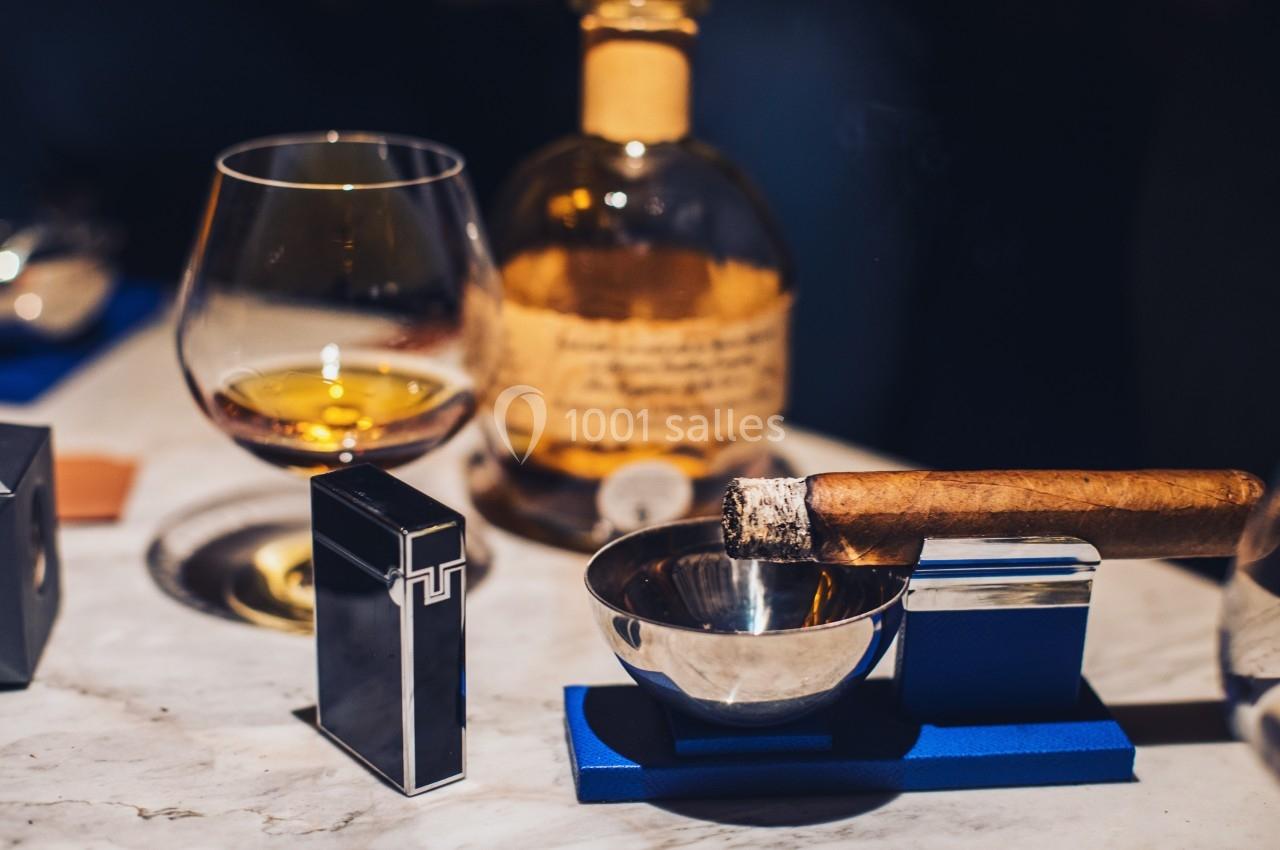Verre de cognac, bouteille en arrière-plan, cigare allumé posé sur un cendrier et briquet sur une table en marbre.