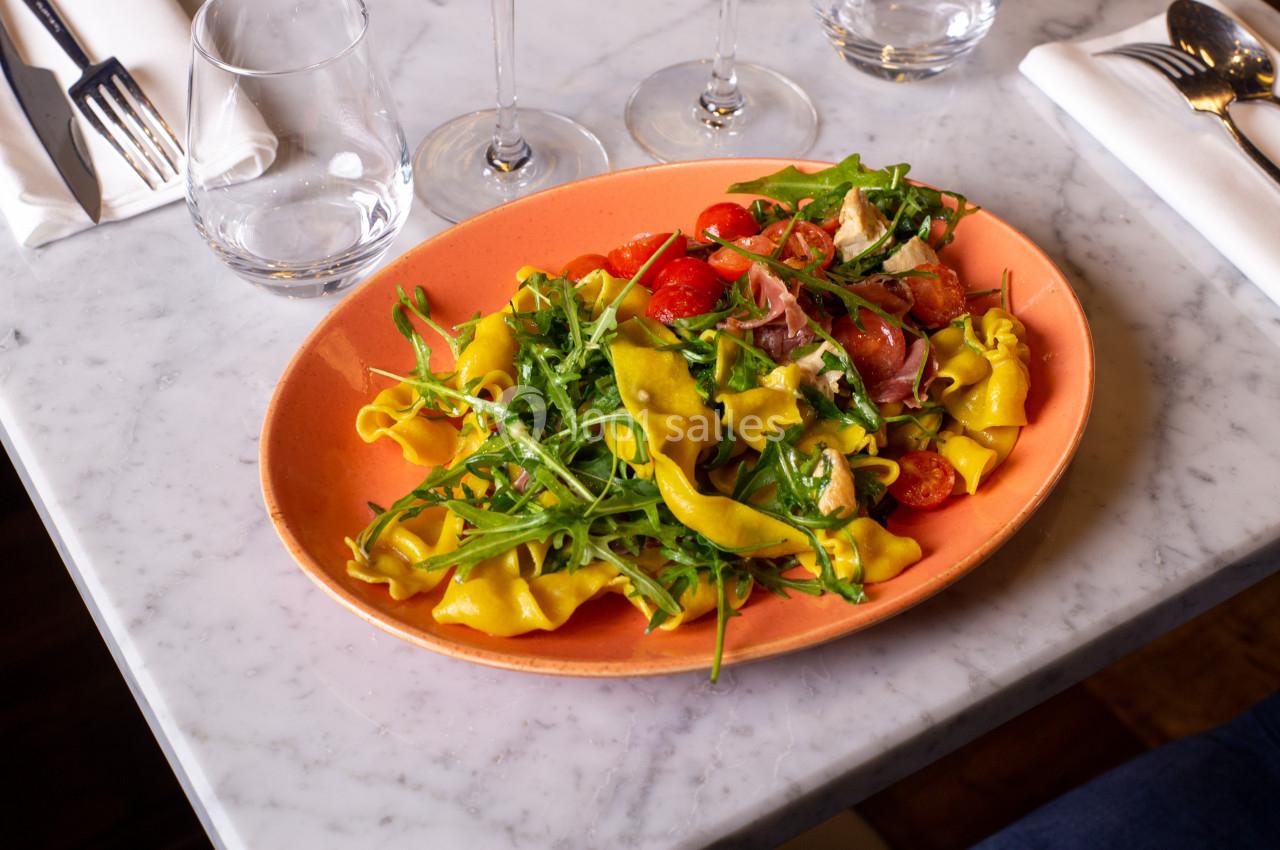 Assiette de pâtes fraîches garnies de roquette, tomates cerises et jambon cru, servie sur une table en marbre.