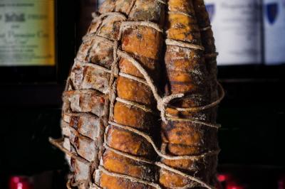 Saucisson sec artisanal suspendu dans un filet devant des bouteilles de vin en arrière-plan flou.