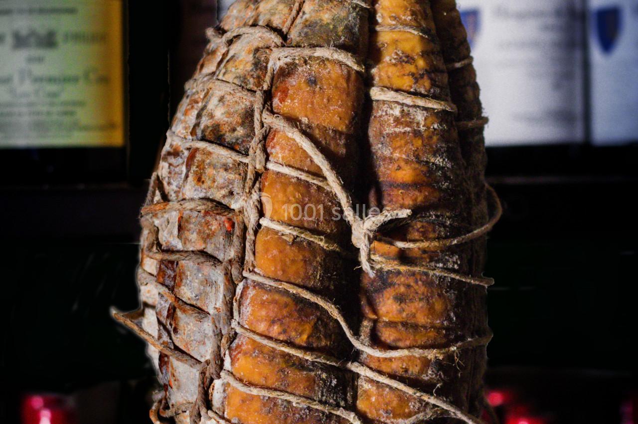 Saucisson sec artisanal suspendu dans un filet devant des bouteilles de vin en arrière-plan flou.