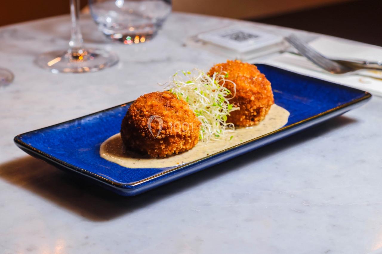 Deux croquettes dorées servies sur une sauce, garnies de pousses vertes, présentées dans une assiette bleue.