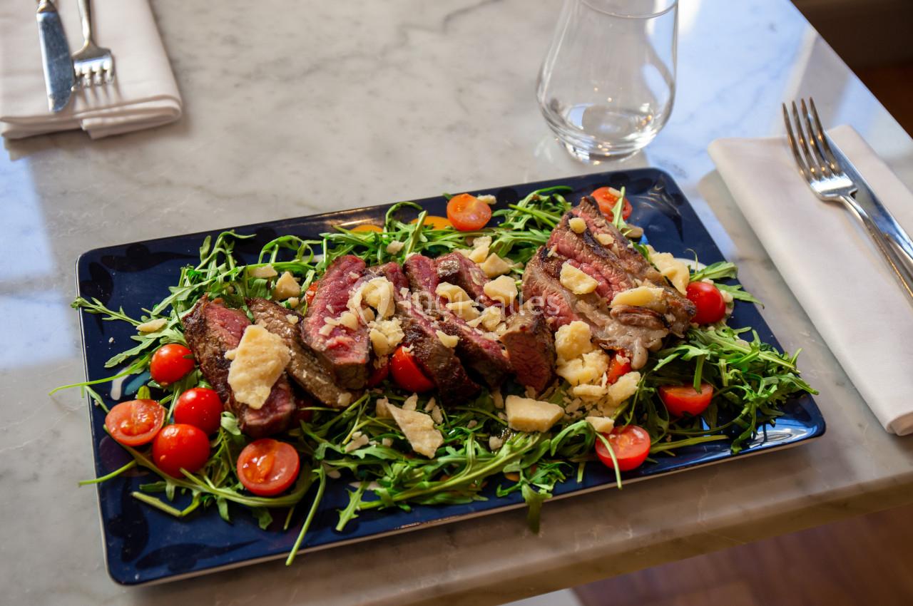 Assiette de viande grillée garnie de roquette, tomates cerises et copeaux de fromage, servie sur une table en marbre.