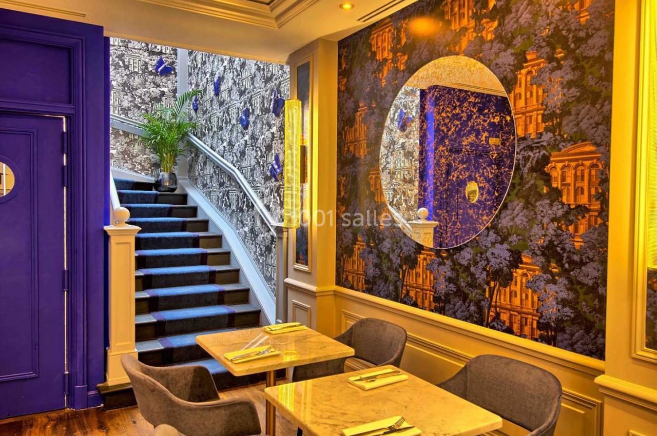 Salle de restaurant colorée avec tables en marbre, chaises grises, escalier bleu et mur décoré de motifs floraux et…
