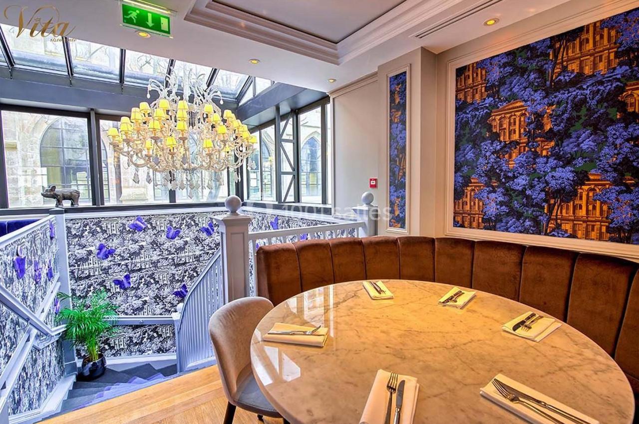 Salle de restaurant élégante avec table ronde, lustre imposant et escalier décoré de motifs floraux colorés.