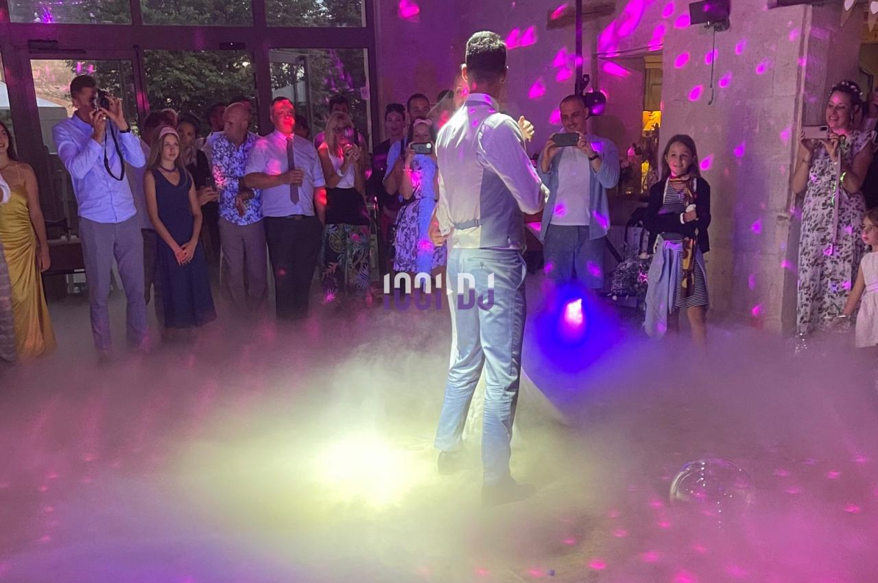 Un homme en costume danse au centre d'une piste entourée d'invités, éclairée par des lumières colorées et de la fumée.