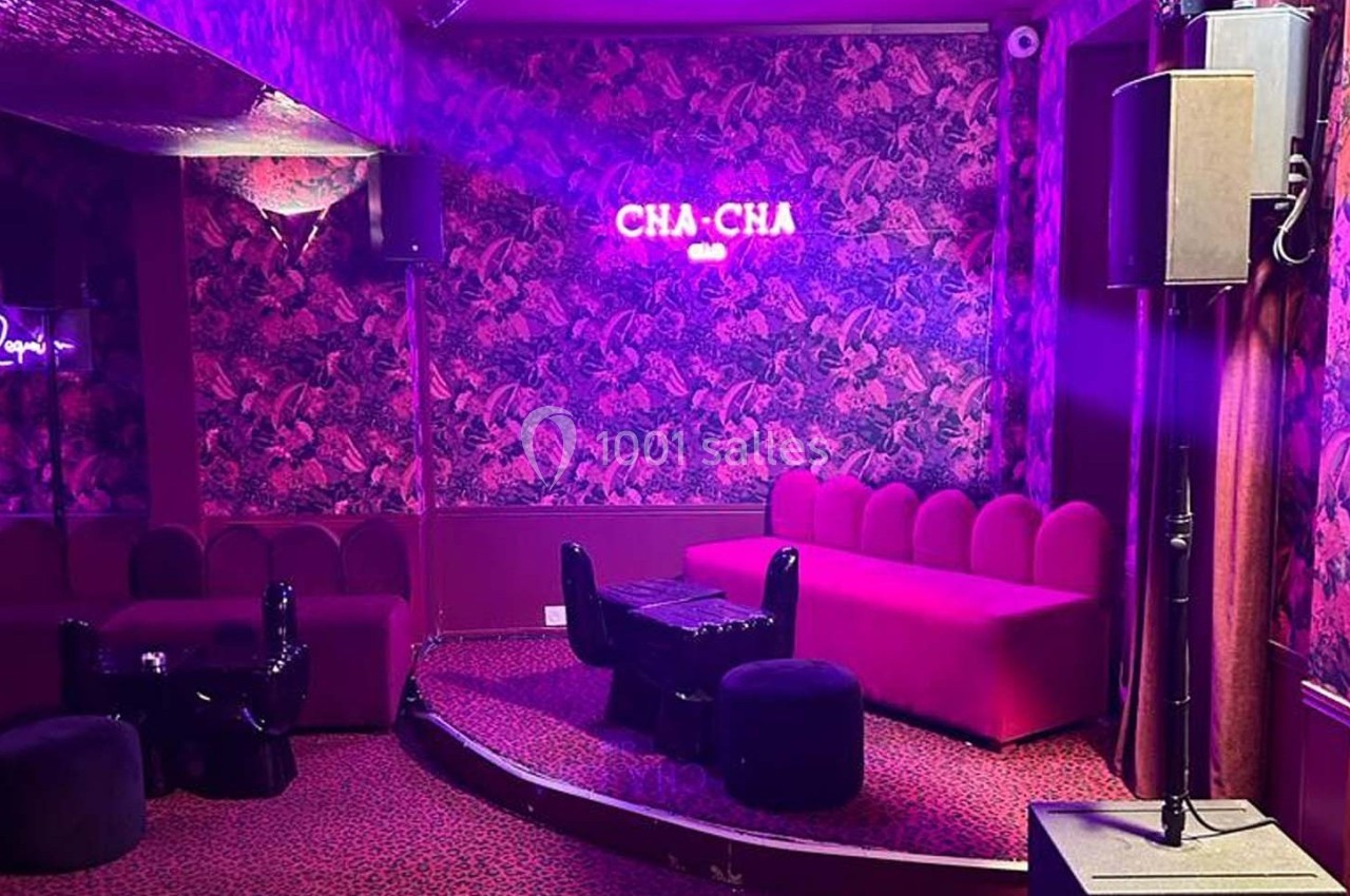 Salle colorée avec canapé rose, murs à motifs, éclairage violet et enseigne lumineuse ’CHA-CHA’.