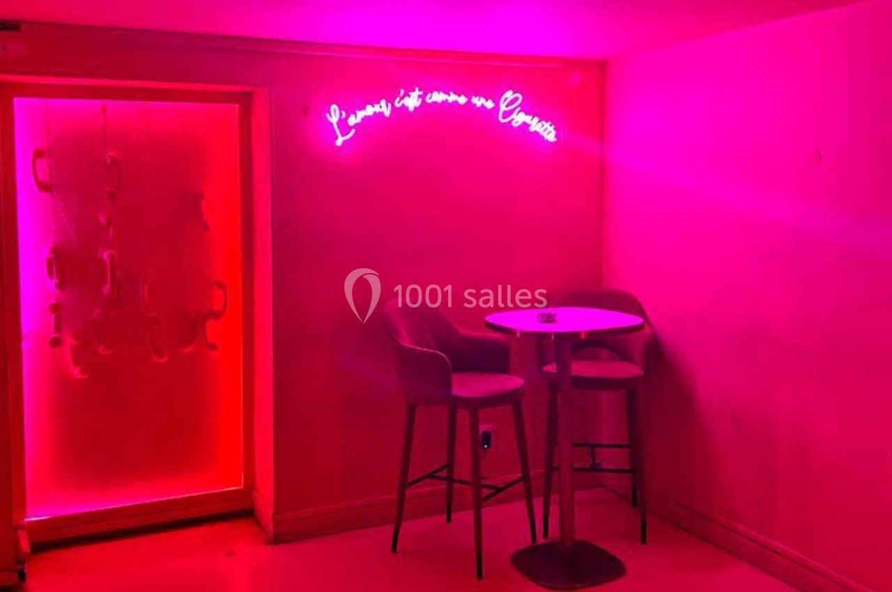 Deux chaises hautes et une table ronde dans une pièce éclairée par une lumière rose avec un néon au mur.