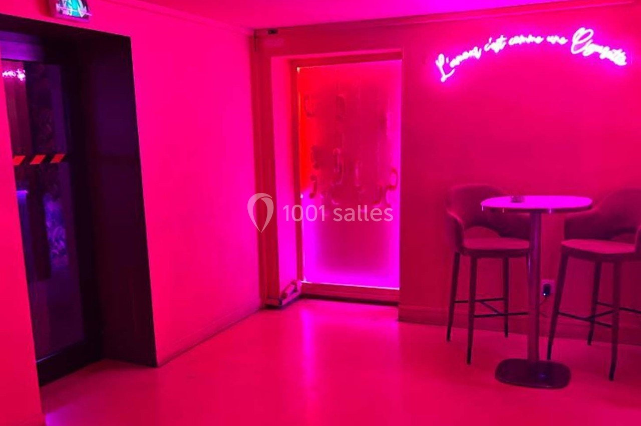 Salle éclairée par une lumière rose avec un néon au mur, une table haute et deux chaises près d'une porte vitrée.