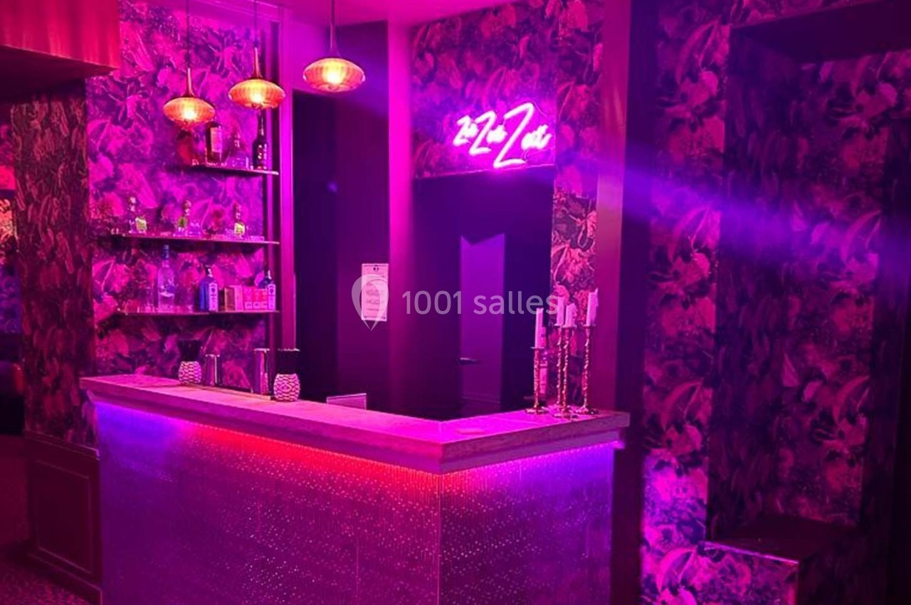 Comptoir de bar éclairé par des lumières roses et violettes, avec décor floral et enseigne néon au mur.