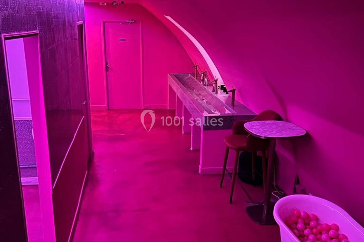 Couloir intérieur aux murs et sol roses, avec un lavabo, une table, une chaise et une bassine remplie de balles.