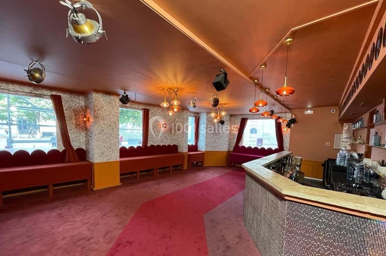 Salle avec moquette rouge, banquettes le long des murs, bar en angle et luminaires suspendus dans un décor rétro.