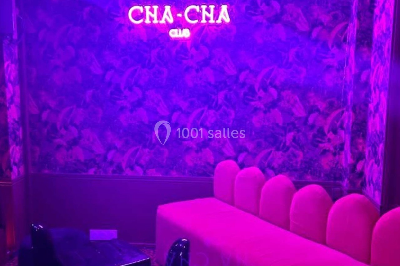 Banquette rose et mur à motifs végétaux éclairés par une lumière violette, avec un néon ’CHA-CHA CLUB’.