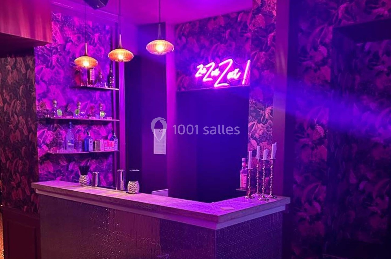 Bar avec comptoir éclairé, étagères de bouteilles, murs décorés et enseigne néon rose dans une ambiance tamisée.