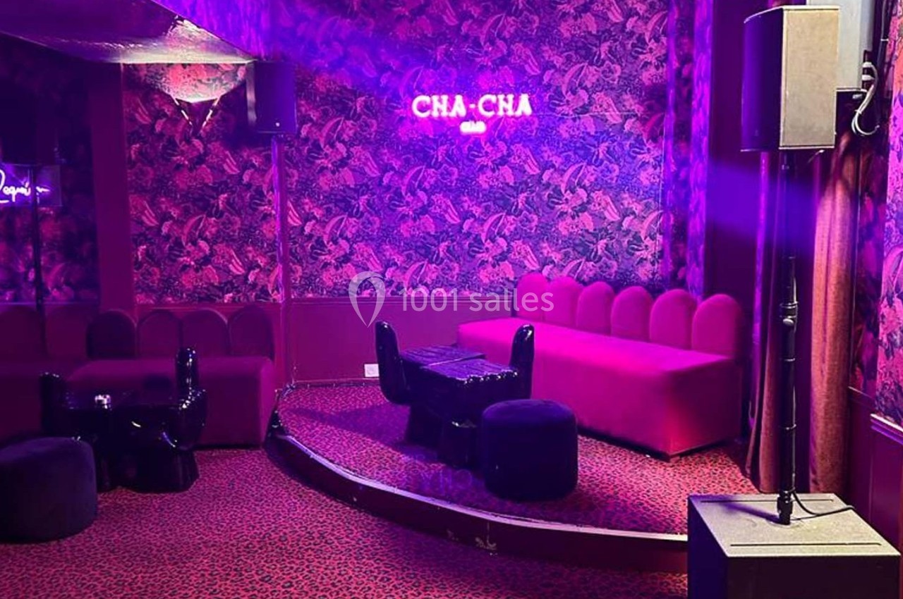 Salle avec banquettes rouges, table basse noire, éclairage tamisé et mur à motif floral, enseigne ’CHA-CHA’ lumineuse.