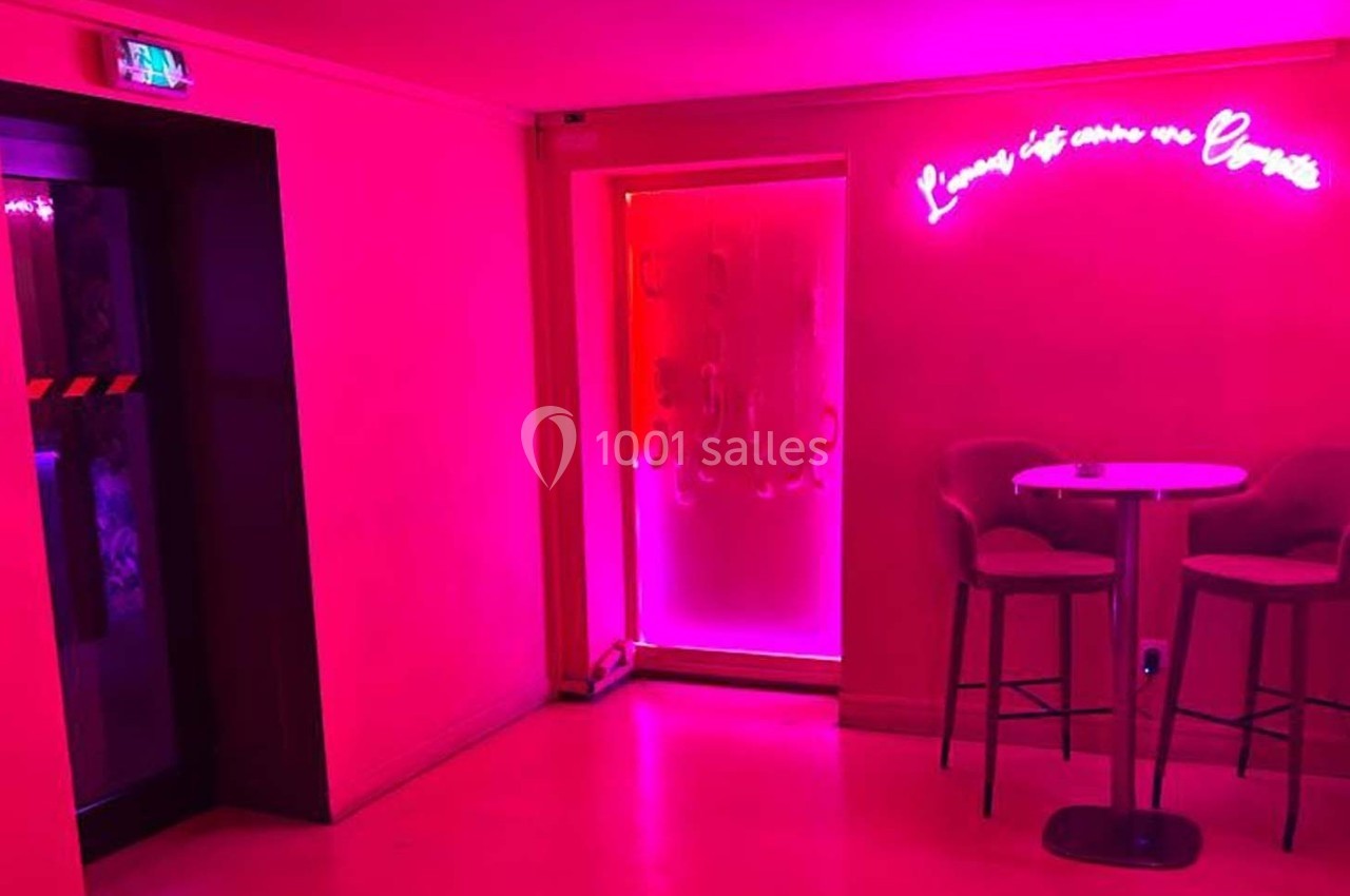 Espace intérieur avec éclairage rose, table haute et chaises, porte vitrée et enseigne lumineuse au mur.