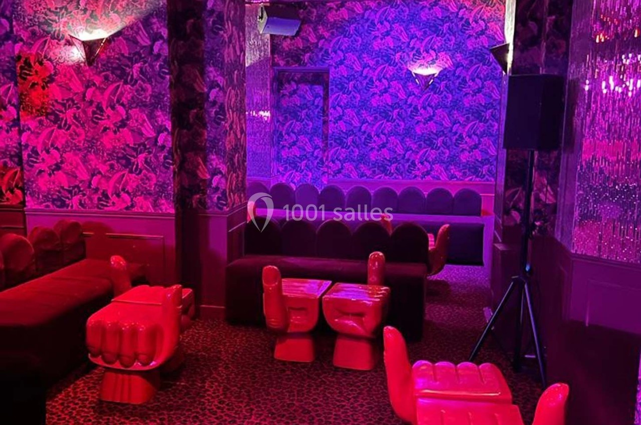 Salle au décor rétro avec fauteuils en forme de mains rouges, murs à motifs et éclairage rose-violet tamisé.