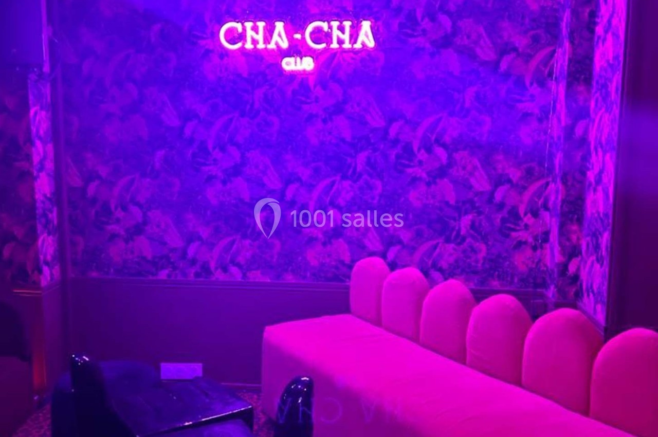 Salle avec un canapé rose, un mur à motifs végétaux et une enseigne néon rose indiquant ’Cha-Cha Club’.