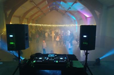 Un DJ en chemise blanche mixe de la musique sur une console lors d'un événement avec des invités en arrière-plan.