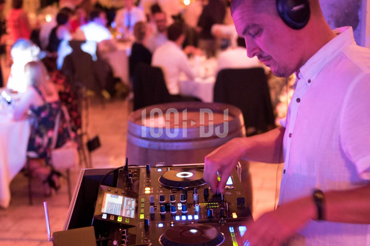 Un DJ en chemise blanche mixe de la musique sur une console lors d'un événement avec des invités en arrière-plan.
