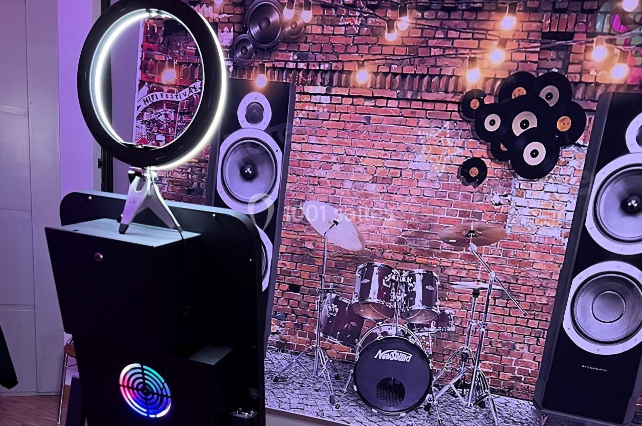 Borne photo avec anneau lumineux devant un décor de mur en briques, batterie et enceintes.