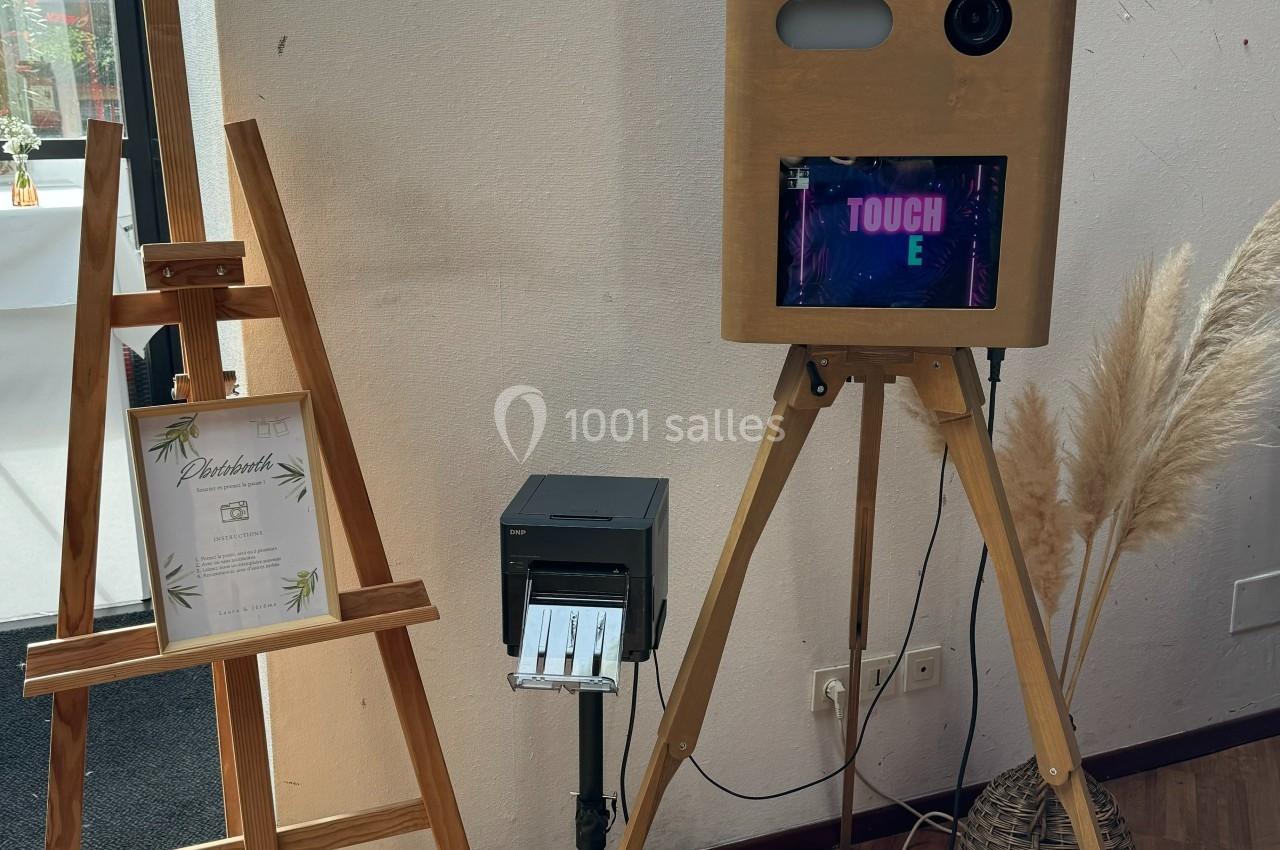 Borne photo en bois sur trépied avec écran tactile, imprimante et anneau lumineux, près d'un chevalet et de décorations.