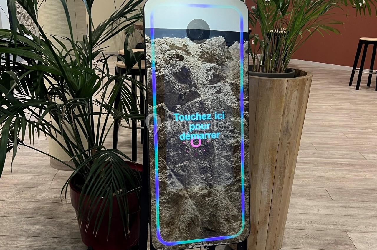 Borne photo interactive avec écran tactile et anneau lumineux, installée dans un espace intérieur avec des plantes…