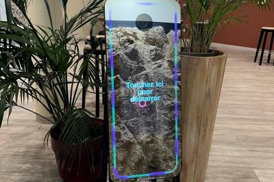 Borne photo interactive avec écran tactile et anneau lumineux, installée dans un espace intérieur avec des plantes…